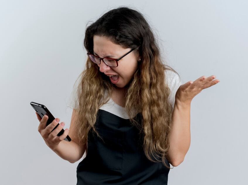 Mujer joven peluquera profesional en delantal mirando la pantalla de su teléfono inteligente volviéndose loca gritando con expresión agresiva de pie sobre la pared blanca